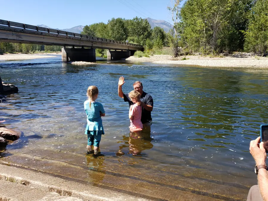 A child being baptized in the Bitterroot River with mountains and a bridge behind